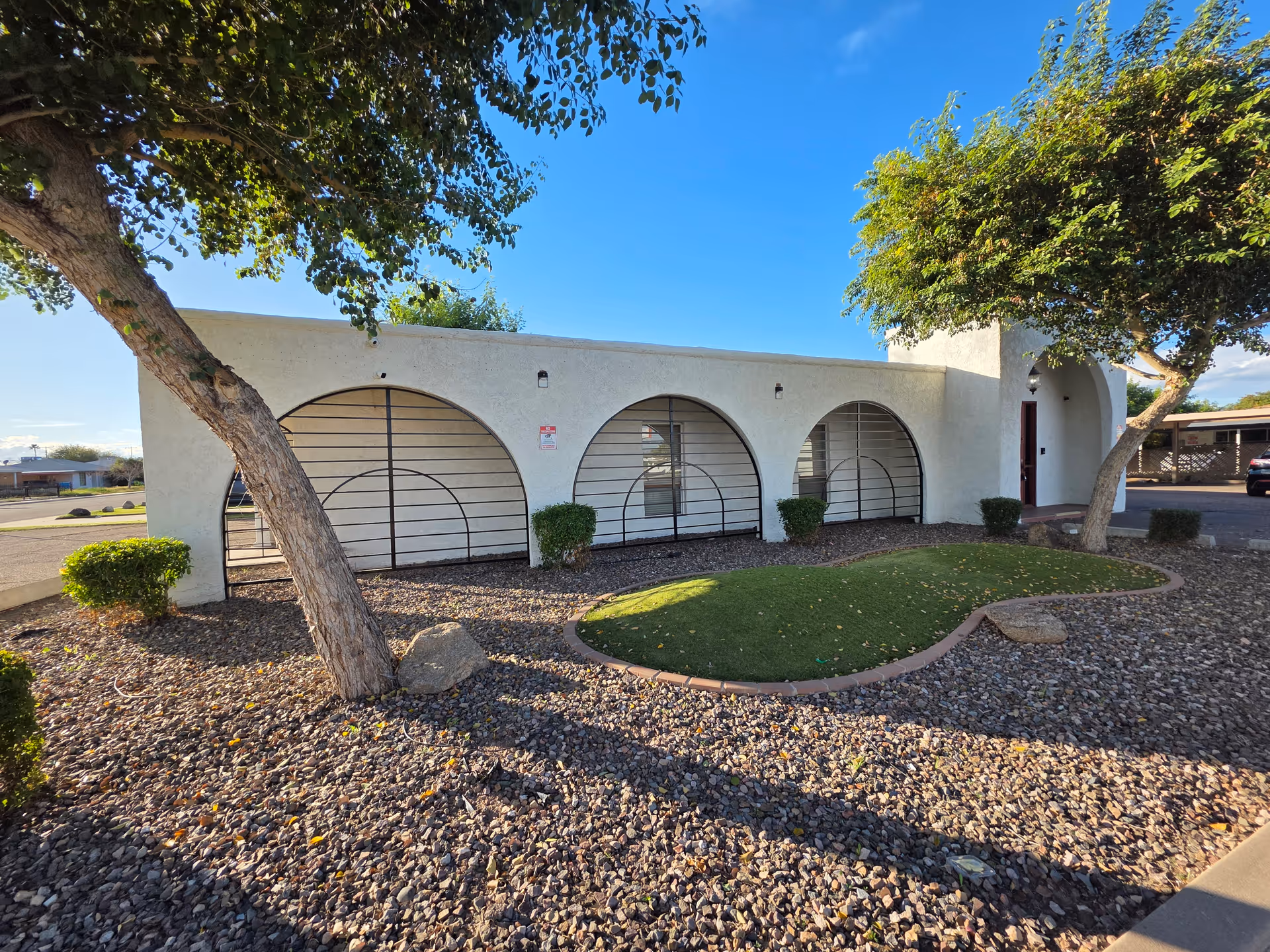 Exterior view of a white building with arched openings, gravel landscaping, and trees.