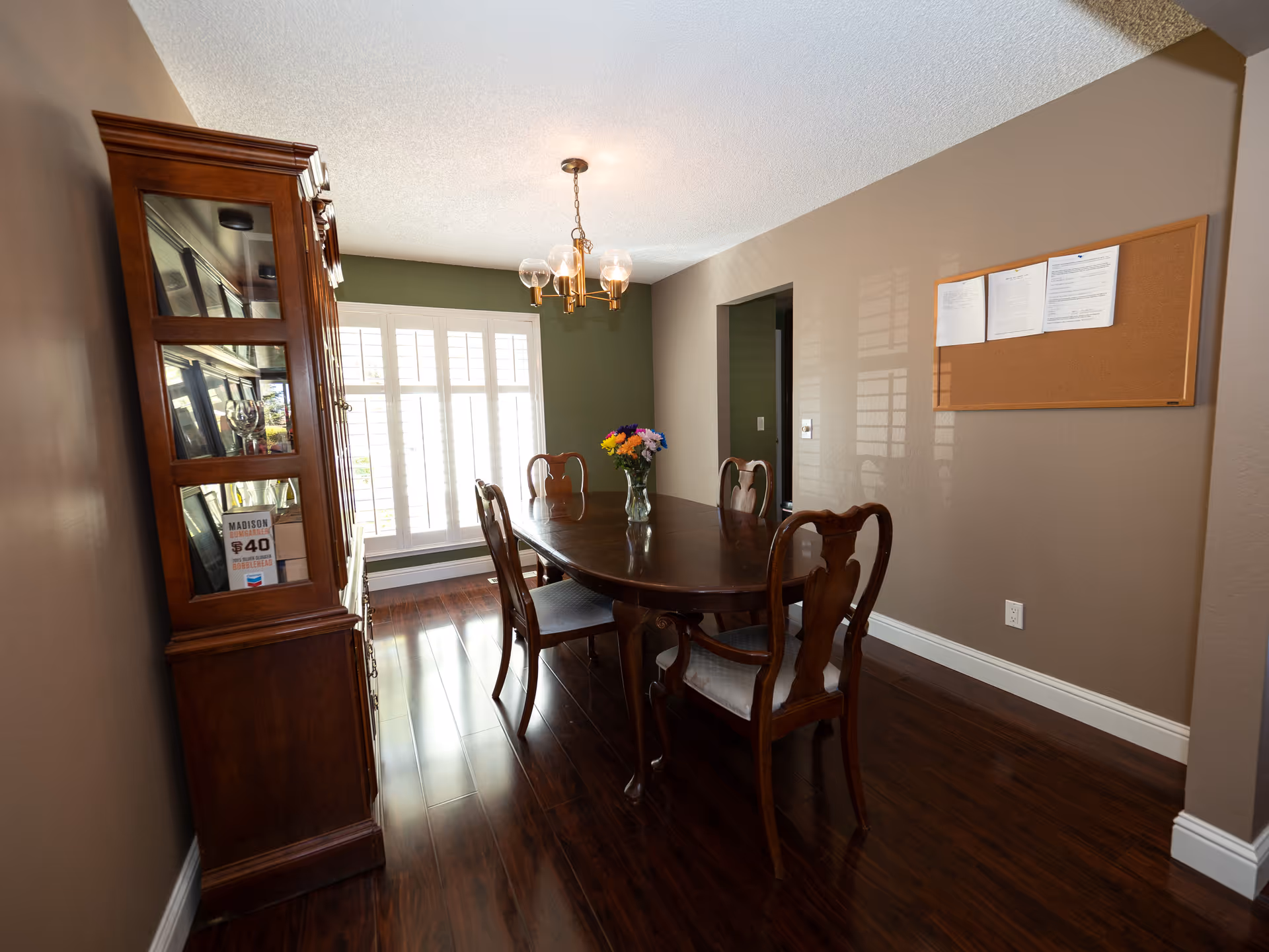 A dining room with a wooden dining table and six matching chairs. A vase with colorful flowers is placed on the table. There is a wooden cabinet with glass doors on the left side and a cork bulletin board with papers pinned on the right wall. The room has wooden flooring and a window with white shutters letting in natural light. A modern chandelier with glass globes hangs from the ceiling.