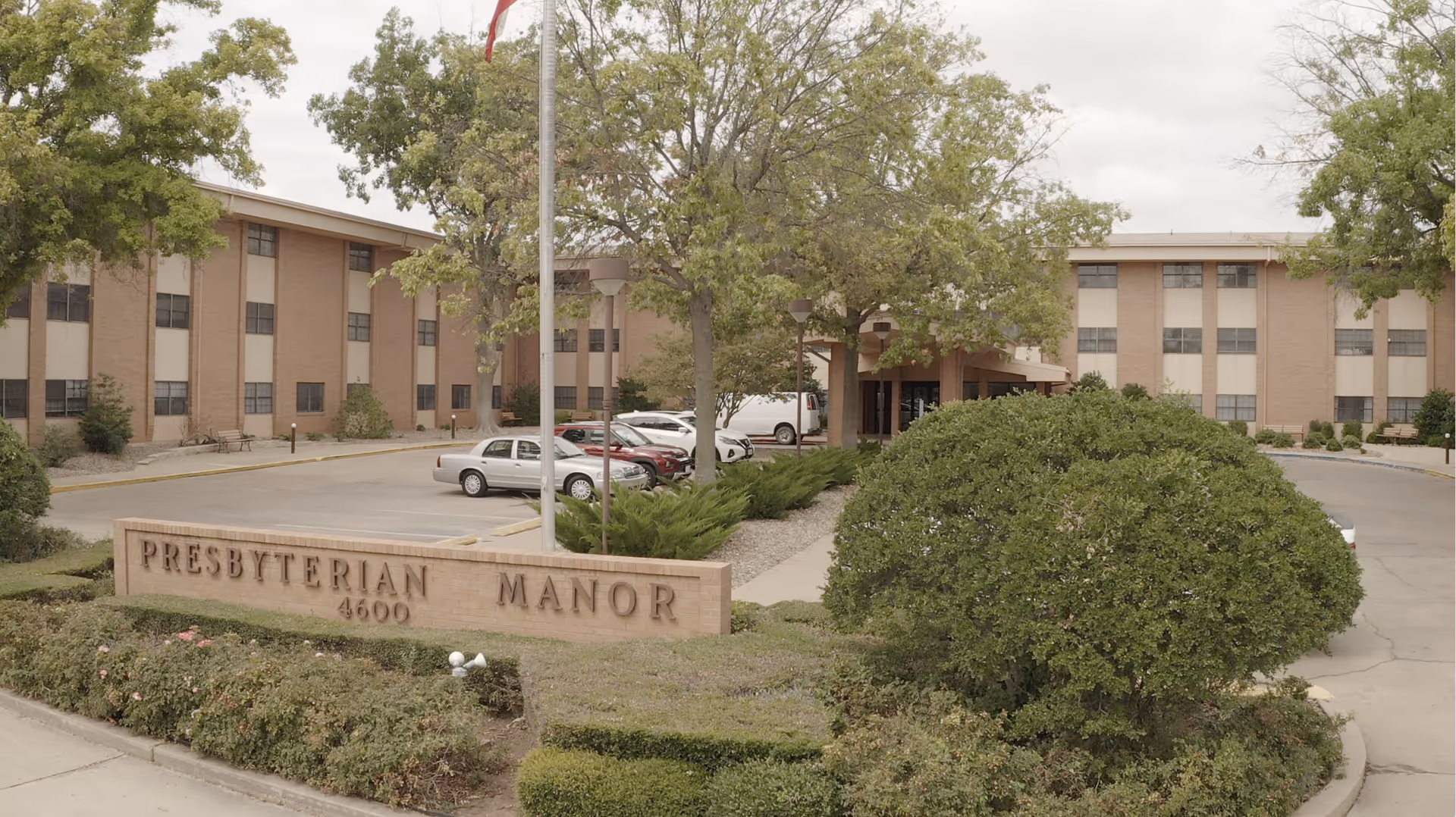 Front view of the Presbyterian Manor building with its sign, entrance canopy, parking lot, flagpole, and landscaping.