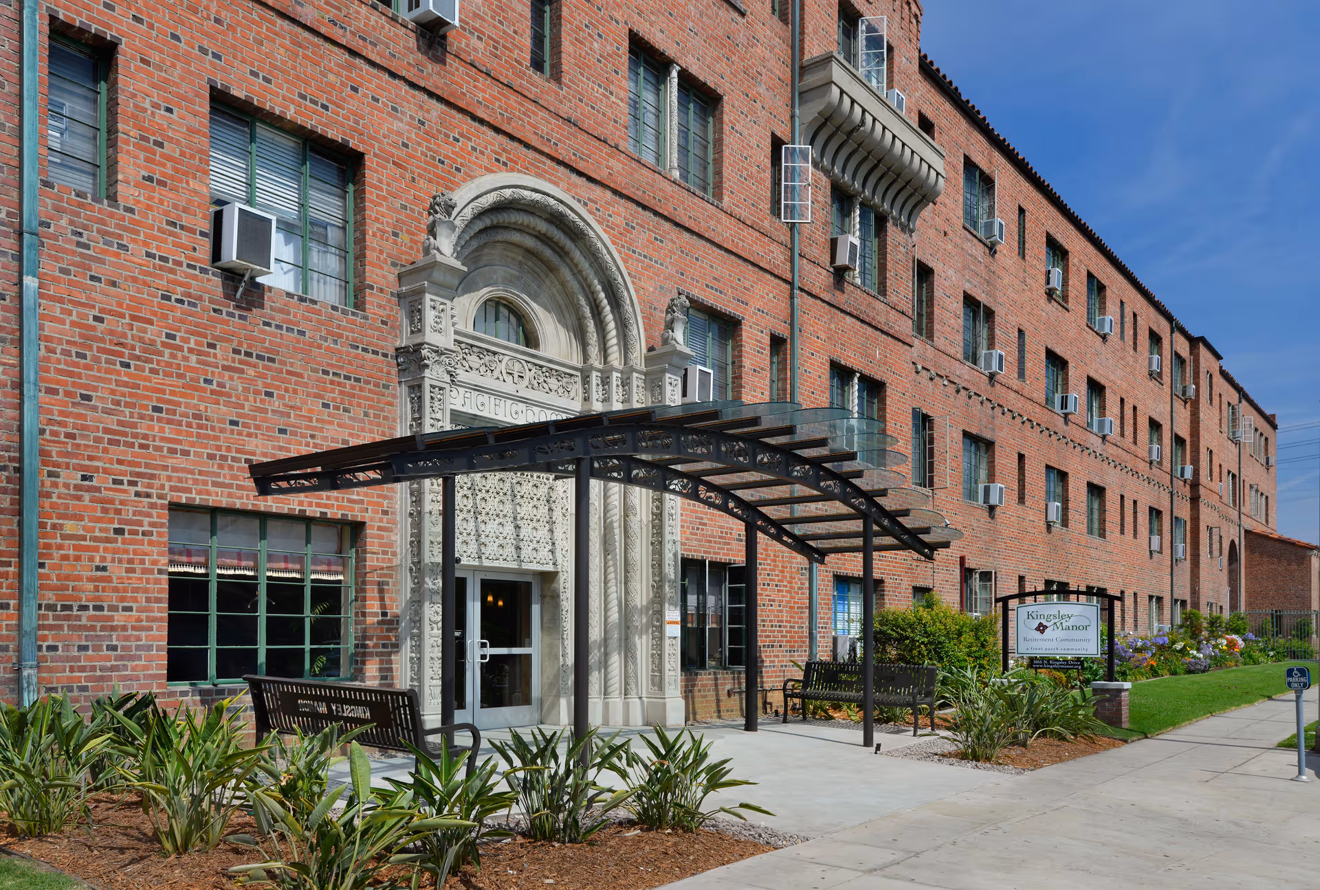 Brick senior living facility exterior with arched entrance, glass canopy, benches, and landscaped walkway.