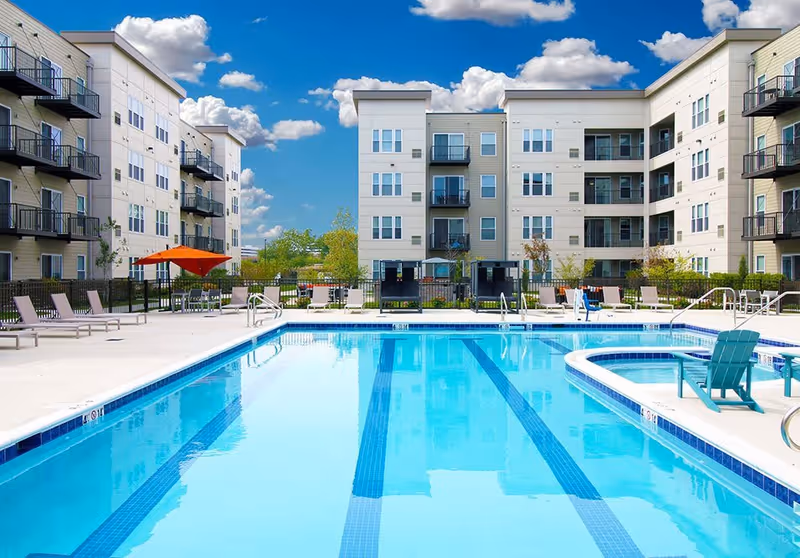 Outdoor swimming pool with clear blue water surrounded by lounge chairs and tables with umbrellas, set in front of a modern multi-story apartment building under a blue sky with scattered clouds.