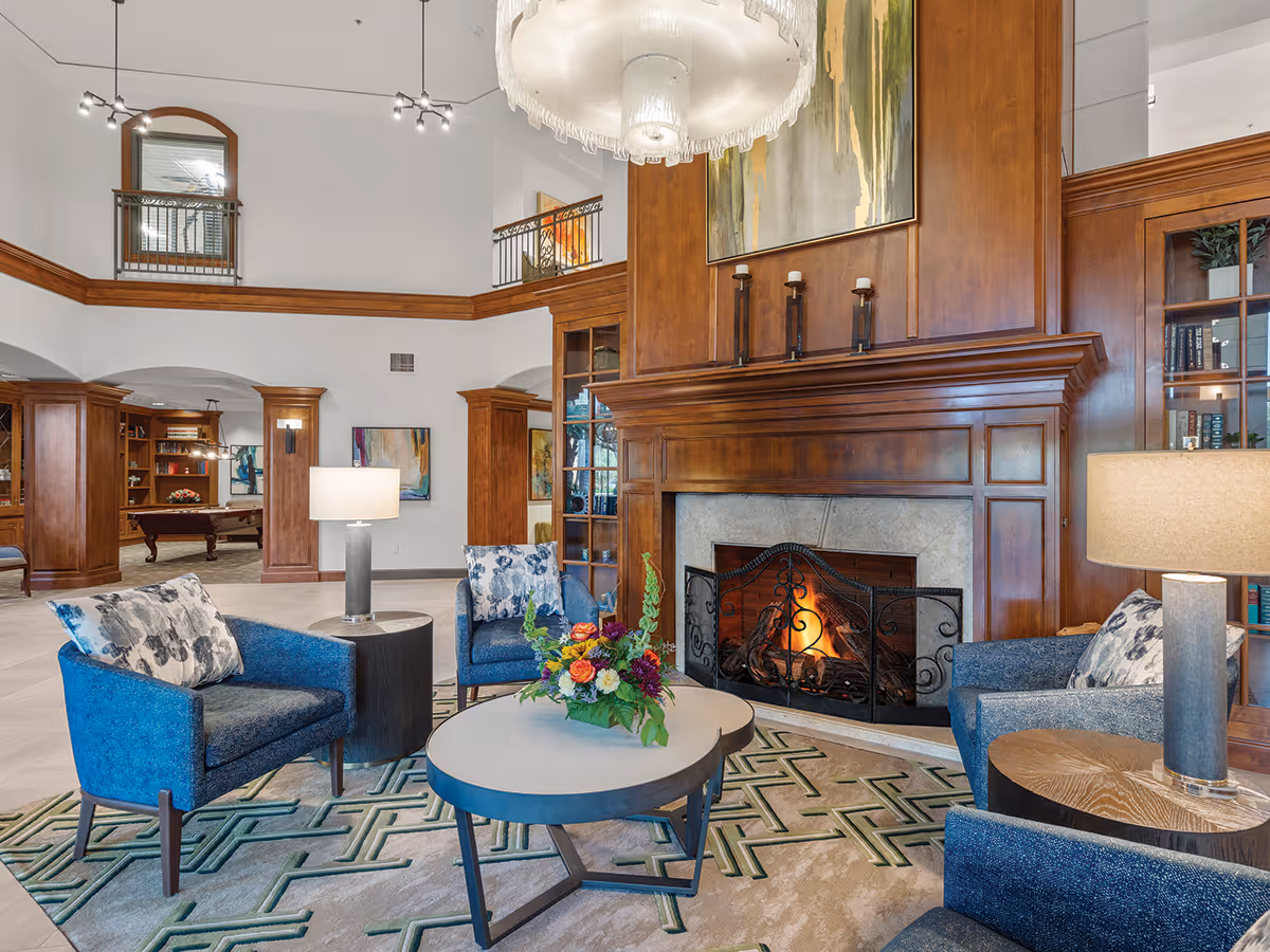 A cozy living room area with a lit fireplace surrounded by wooden paneling. The room features three blue armchairs with patterned cushions, two side tables with lamps, and a round coffee table with a floral arrangement. The space has a high ceiling with a large chandelier and an open view into an adjacent room with a pool table and bookshelves.