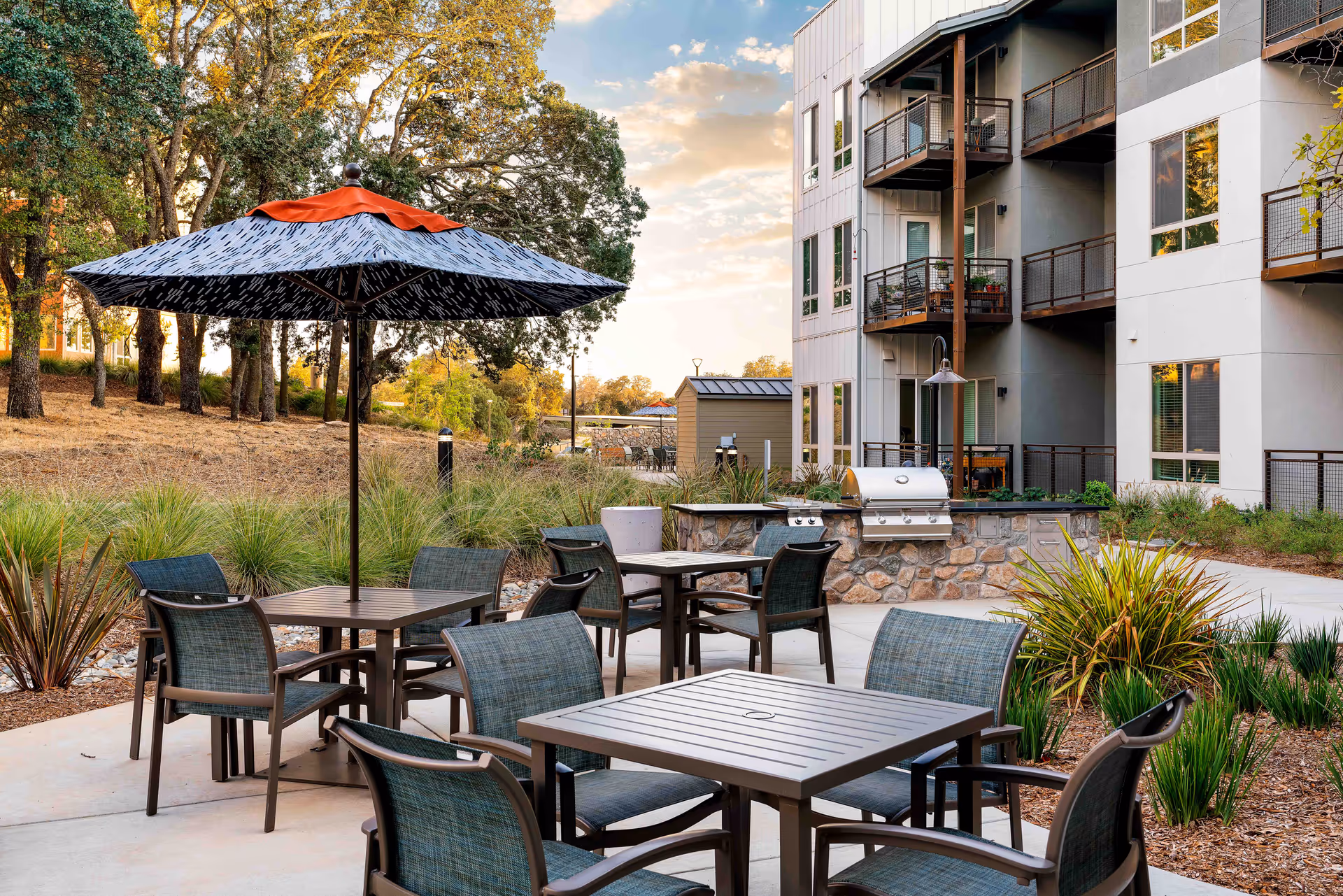 Outdoor patio with tables, chairs and a large umbrella next to a barbecue area and a multi-story residential building.