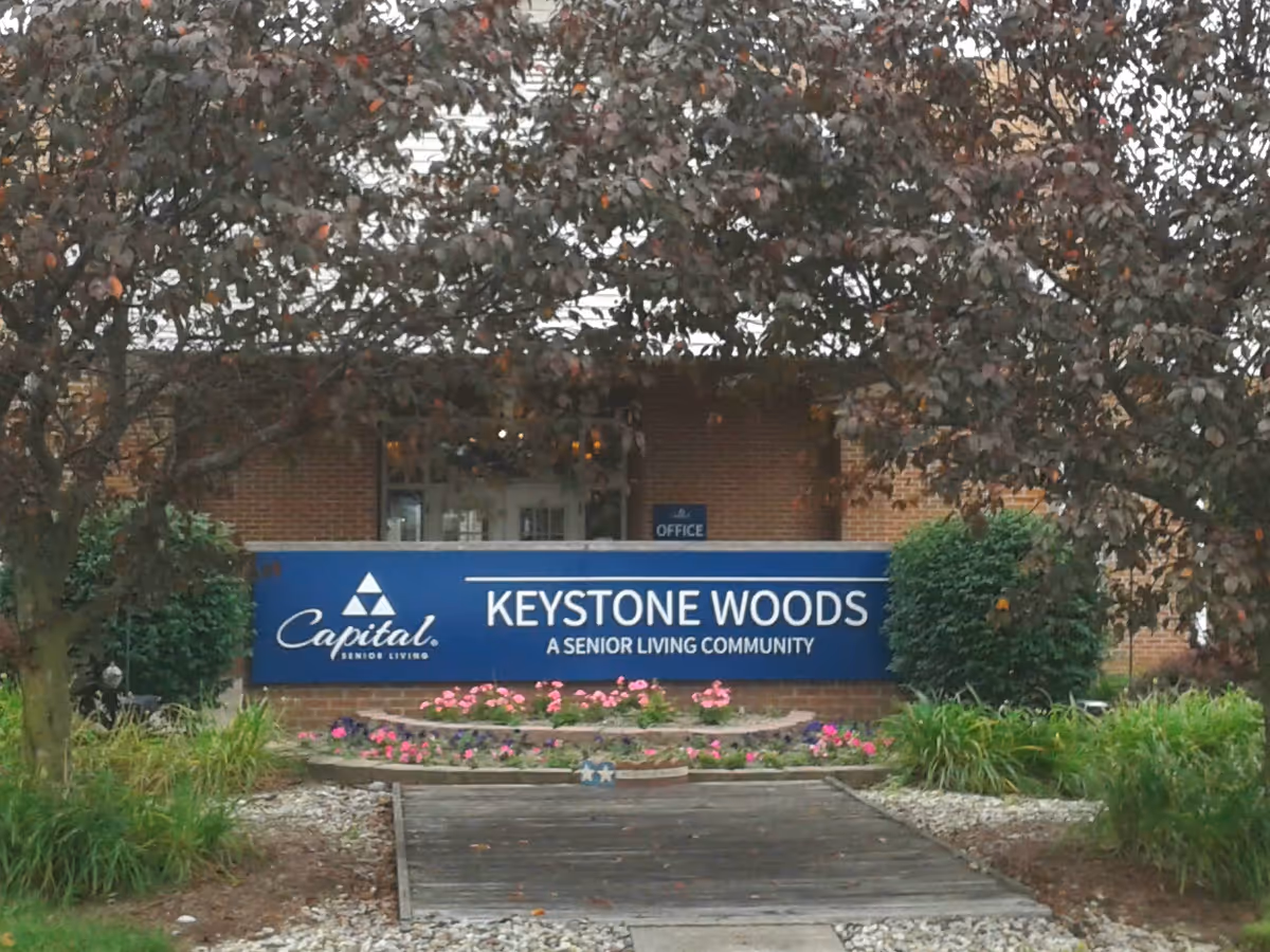 Exterior view of Keystone Woods senior living sign with plants and a brick building behind.