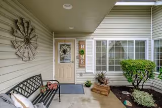 Covered front porch area of a residential building with a beige door decorated with a wreath, a vertical wooden sign spelling HOME, a black metal bench with cushions, a decorative metal wall piece, potted plants, and a small landscaped garden with a trimmed bush and mulch.