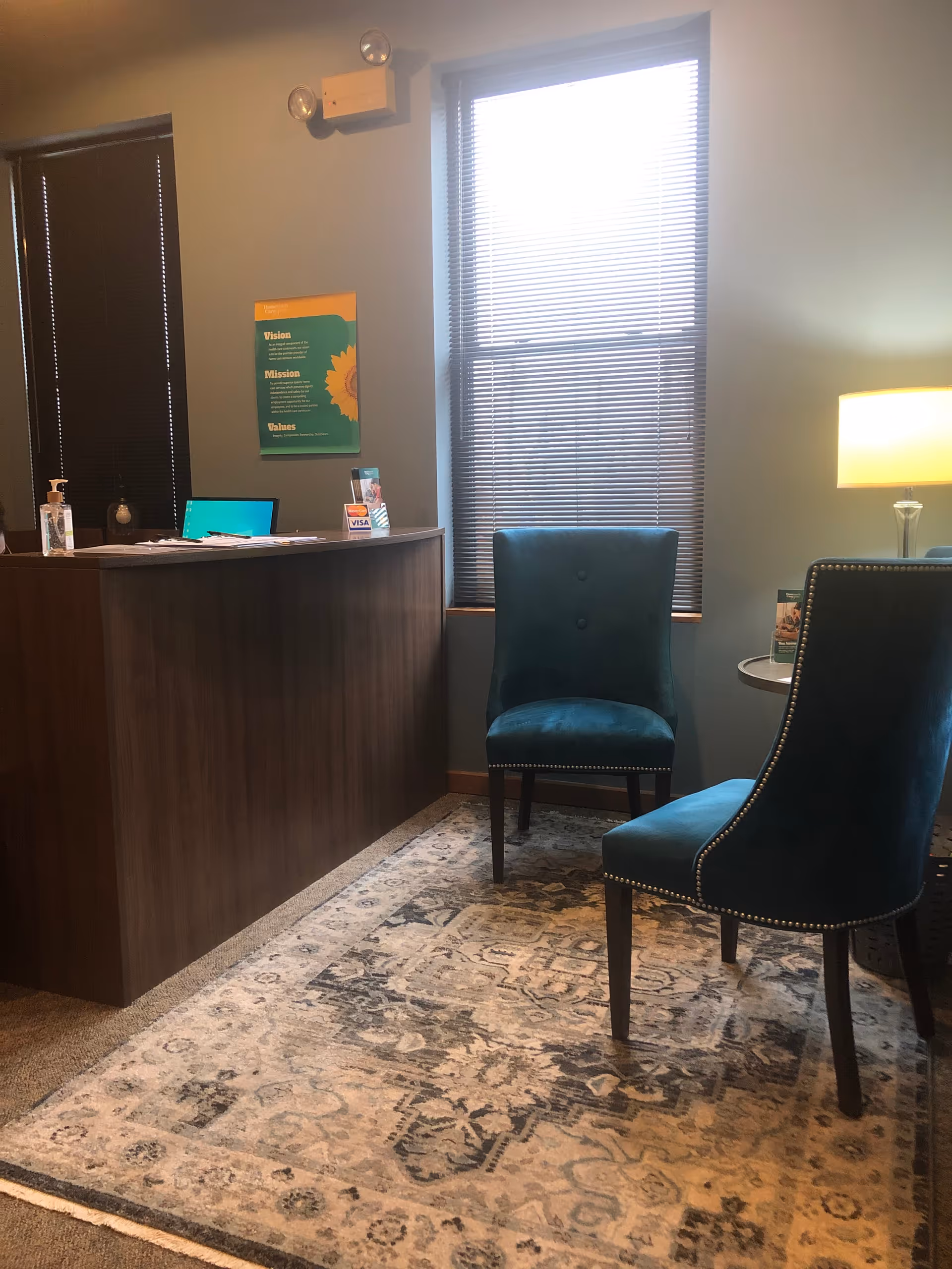 Reception area featuring a dark wood desk, two teal chairs, rug, and large window blinds.