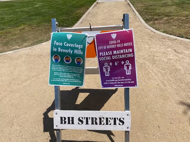 A signpost on a dirt pathway with two signs attached. The left sign is green and instructs people to wear face coverings in Beverly Hills, showing illustrations of people wearing masks. The right sign is purple and reminds people to maintain social distancing of 6 feet, with icons of two people standing apart. Below the signs, a white wooden board reads 'BH STREETS'. The pathway is bordered by grass on both sides.
