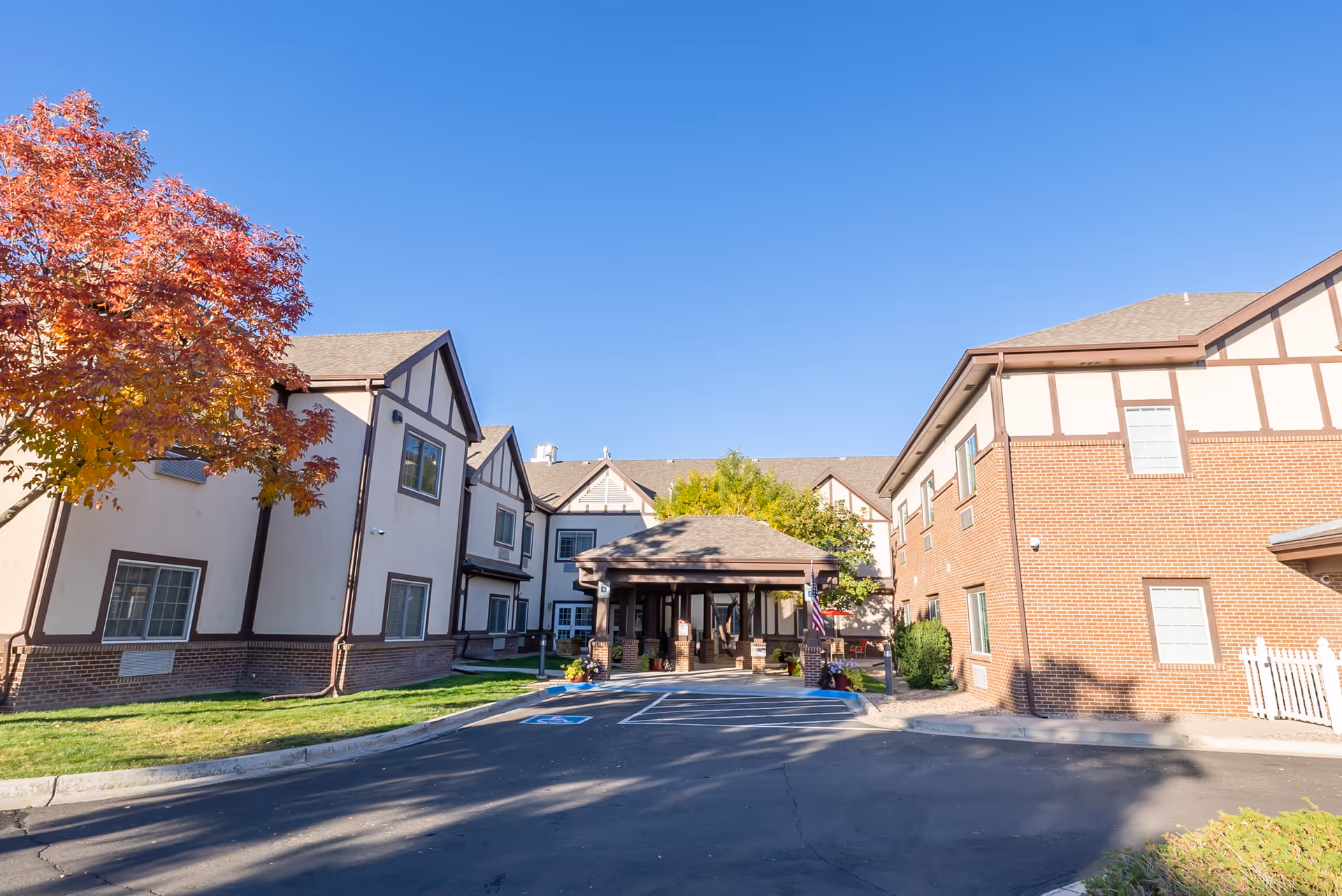 Exterior view of Chateau At Sharmar Assisted Living facility showing a two-story building with a mix of brick and beige siding, a covered entrance, a tree with autumn-colored leaves, and a clear blue sky.