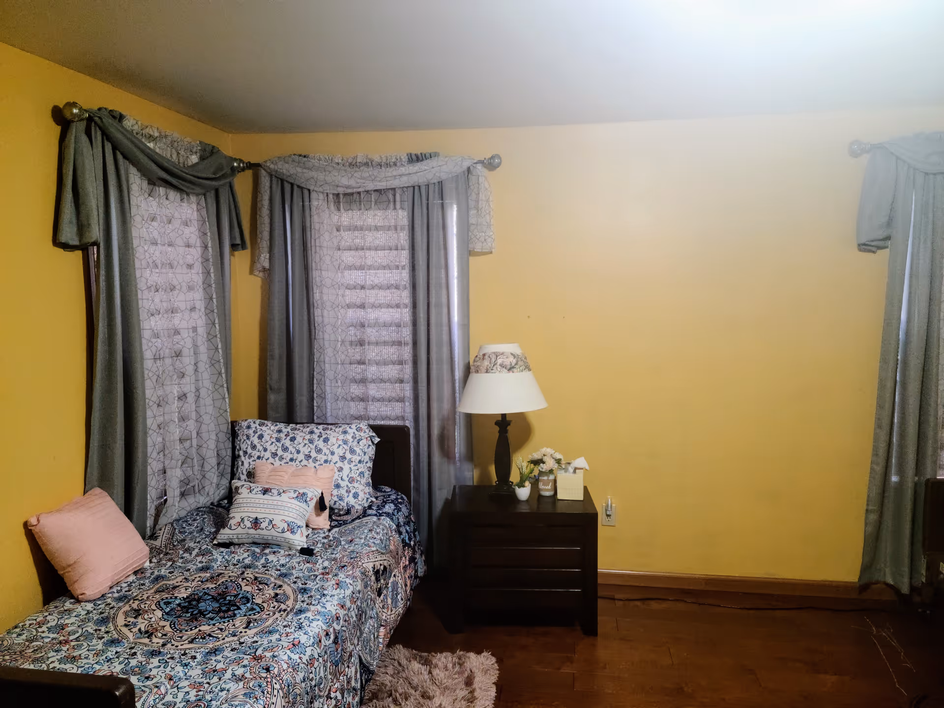 Yellow-walled bedroom with a patterned bed, curtains, nightstand, lamp, and wooden floor.