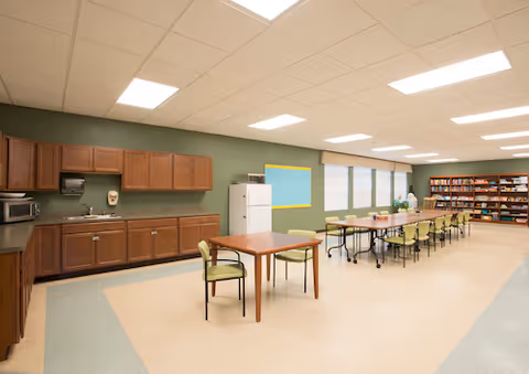 A spacious room with a kitchenette area featuring wooden cabinets, a microwave, a refrigerator, and a paper towel dispenser. There are several tables and chairs arranged for dining or activities, with a long table and multiple chairs along one side. The room has large windows with blinds and a bookshelf filled with books at the far end. The walls are painted green and the ceiling has recessed lighting.