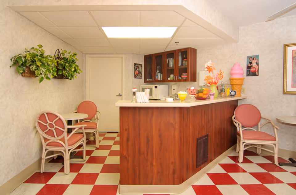 A small indoor dining area with a checkered red and white floor, featuring a wooden counter with various items on top including a large pink ice cream cone decoration, a bowl of fruit, and a glass of yellow beverage. There are three cushioned chairs with light-colored frames and red upholstery around small round tables. Two hanging green plants are mounted on the wall to the left, and there are framed pictures on the walls.