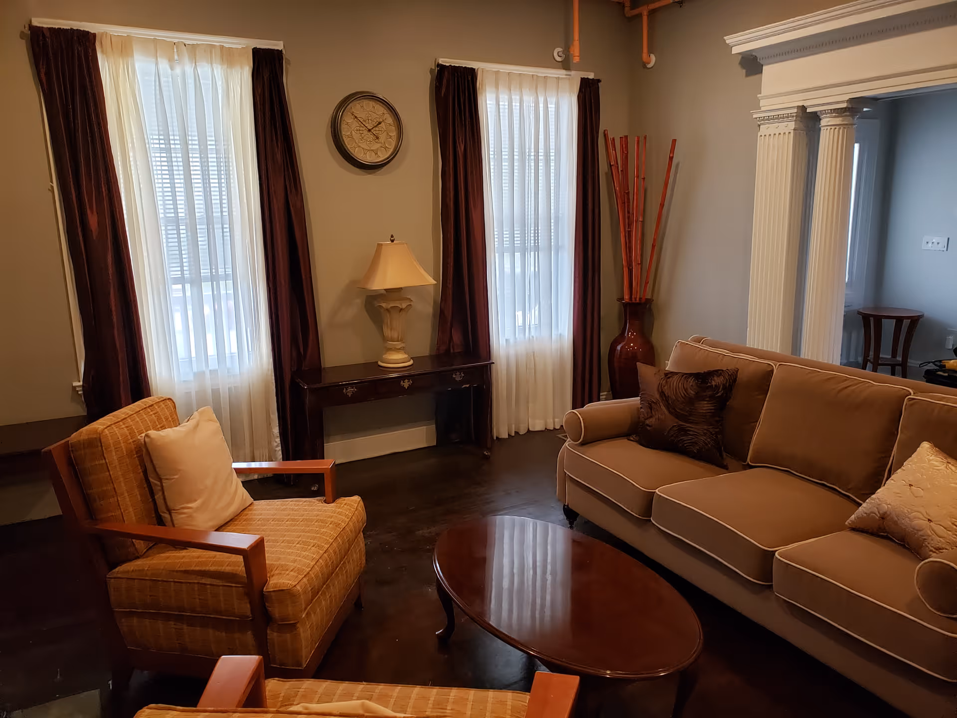 A cozy living room with two armchairs and a sofa with cushions arranged around a wooden oval coffee table. The room has two tall windows with sheer white curtains and dark drapes. A small table with a lamp and a wall clock is positioned between the windows. There is a large decorative vase with tall sticks in the corner and white columns partially visible on the right side.
