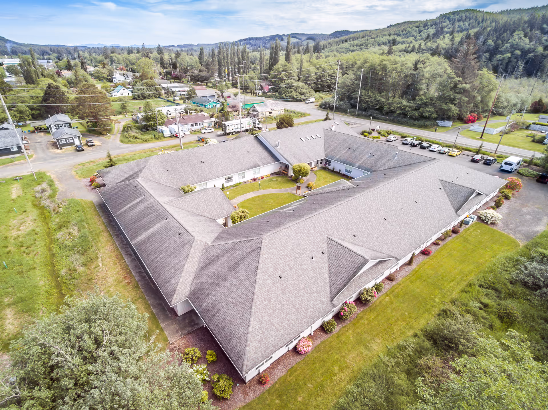 Aerial view of a large, single-story building with a courtyard in the center, surrounded by green lawns and trees. The building is located in a semi-rural area with other houses and roads visible in the background under a partly cloudy sky.