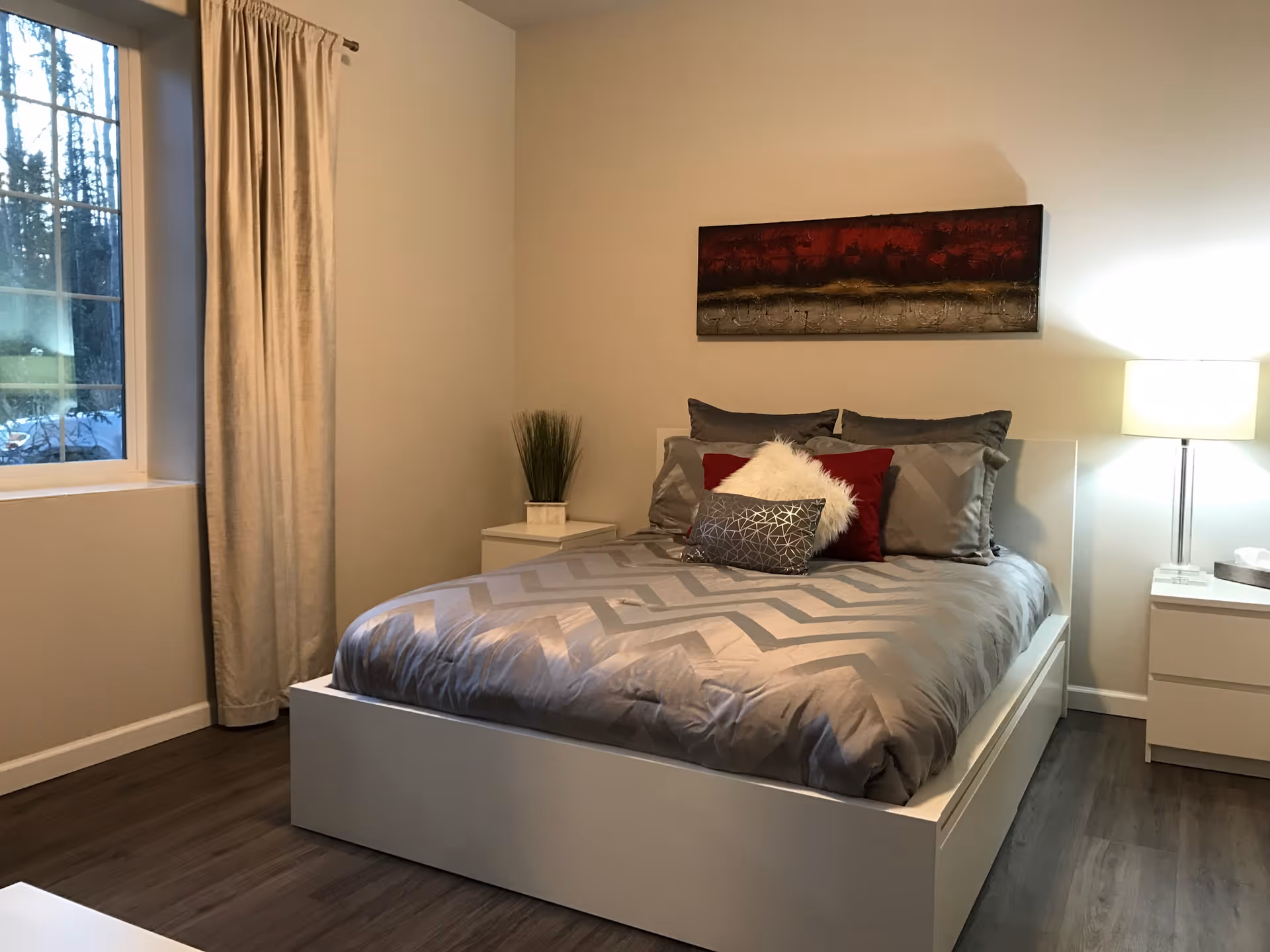 Neatly made bed with gray bedding, decorative pillows, nightstands and a lamp in a modern bedroom with a window and wall art.