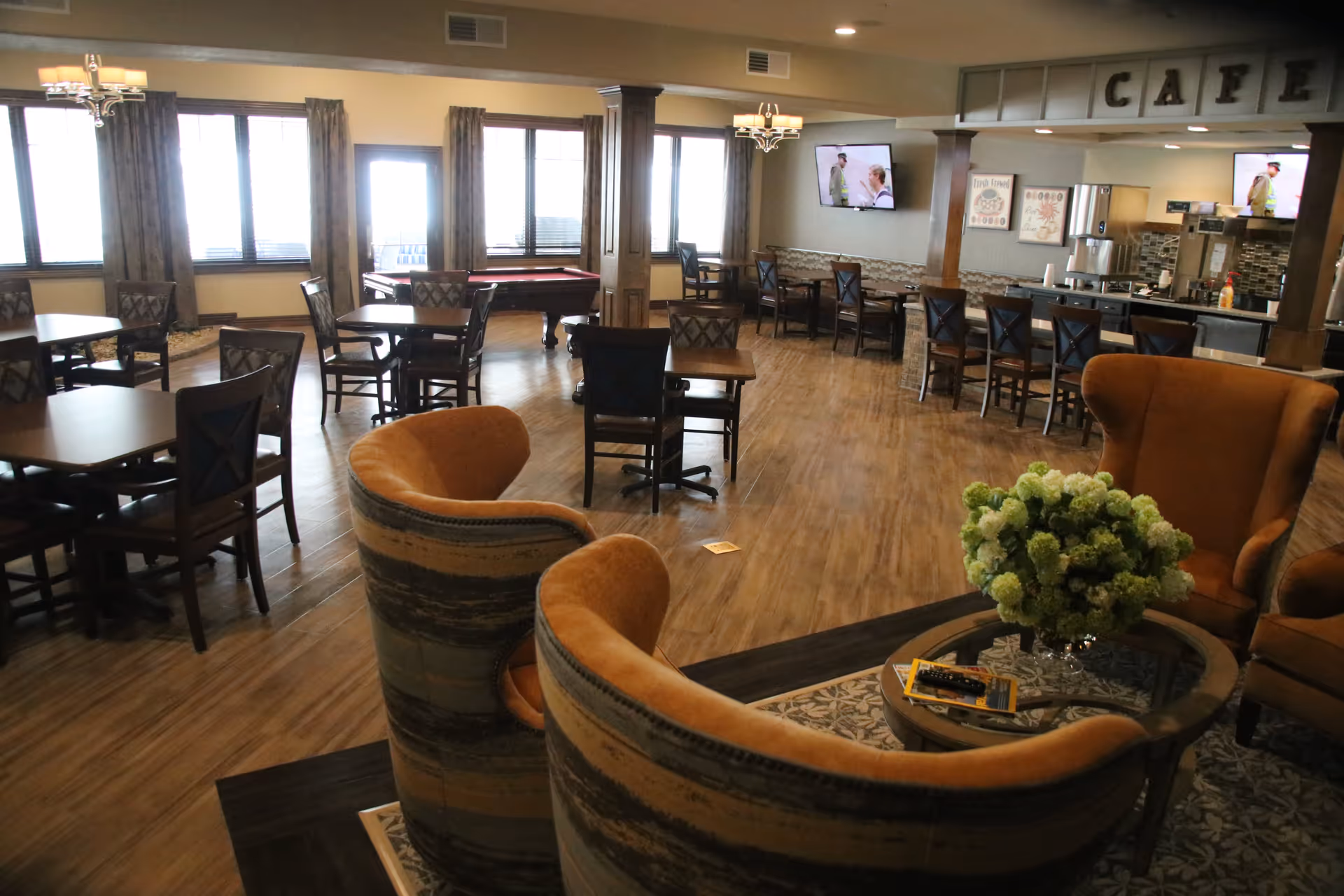 A spacious and well-lit common area with multiple seating options including cushioned armchairs around a glass coffee table with a flower arrangement, several wooden tables with chairs, and a bar counter with high stools. The room features large windows with curtains, two wall-mounted televisions, and a pool table near the windows. The ceiling has chandeliers and the floor is wooden. The area is labeled as a cafe with a sign above the bar counter.