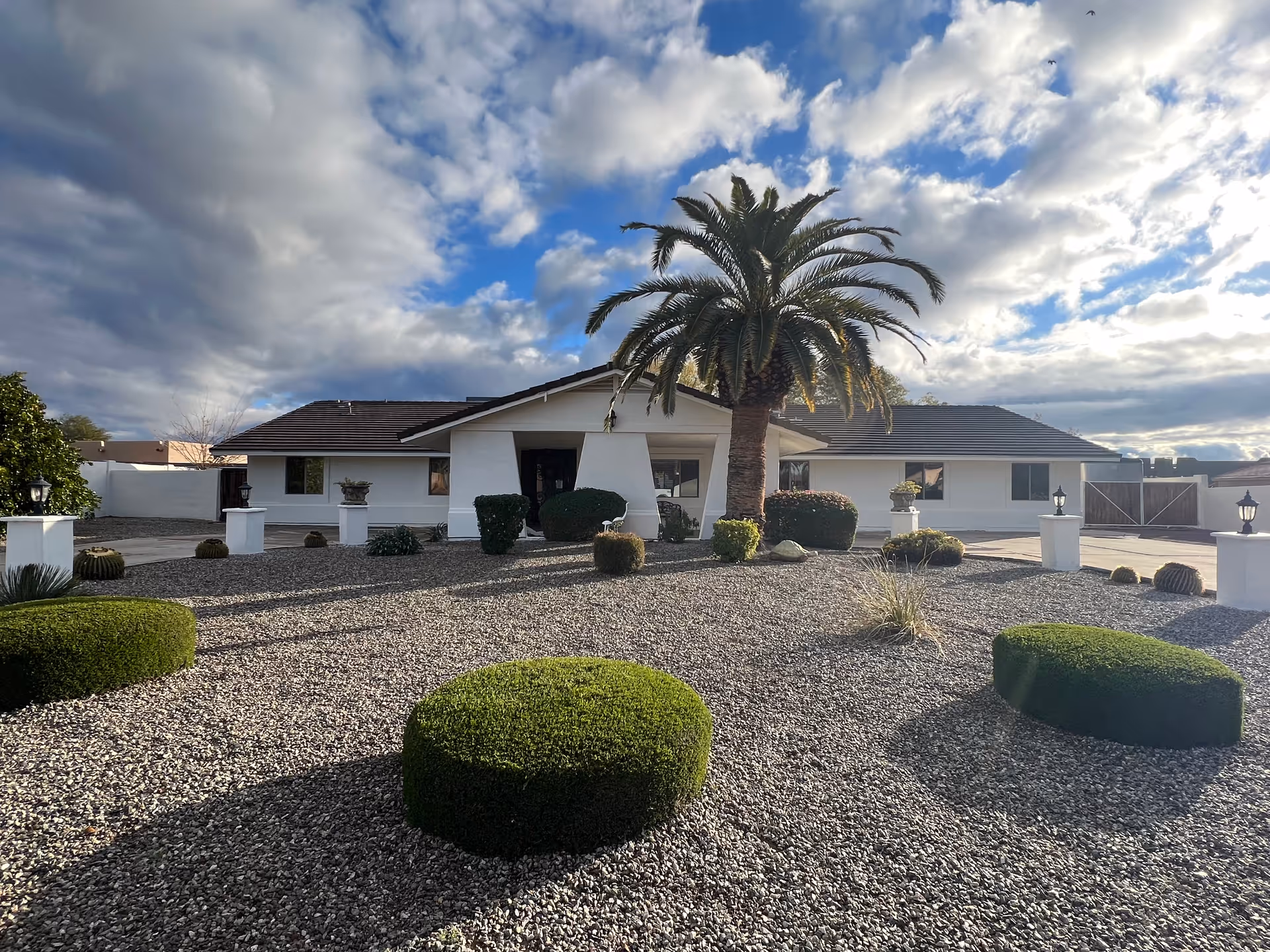 Front exterior view of a single-story white building with a dark roof, surrounded by a landscaped yard featuring gravel, neatly trimmed bushes, a large palm tree in the center, and several white pillars with lanterns. The sky is partly cloudy with patches of blue.
