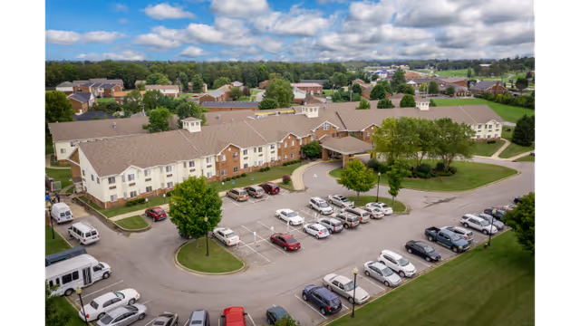 Aerial view of McDowell Place of Danville, a large senior living facility with a two-story building surrounded by a parking lot filled with cars. The building has a beige and brick exterior with multiple windows and cupolas on the roof. The surrounding area includes green lawns, trees, and a partly cloudy sky.