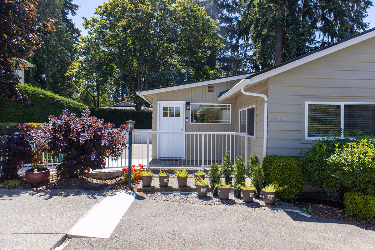 Front view of a single-story beige building with a white door and railing, potted plants.
