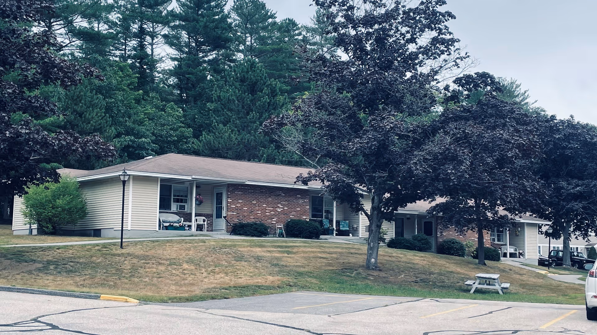 Single-story senior living building with brick facade, lawn, trees, and outdoor seating.