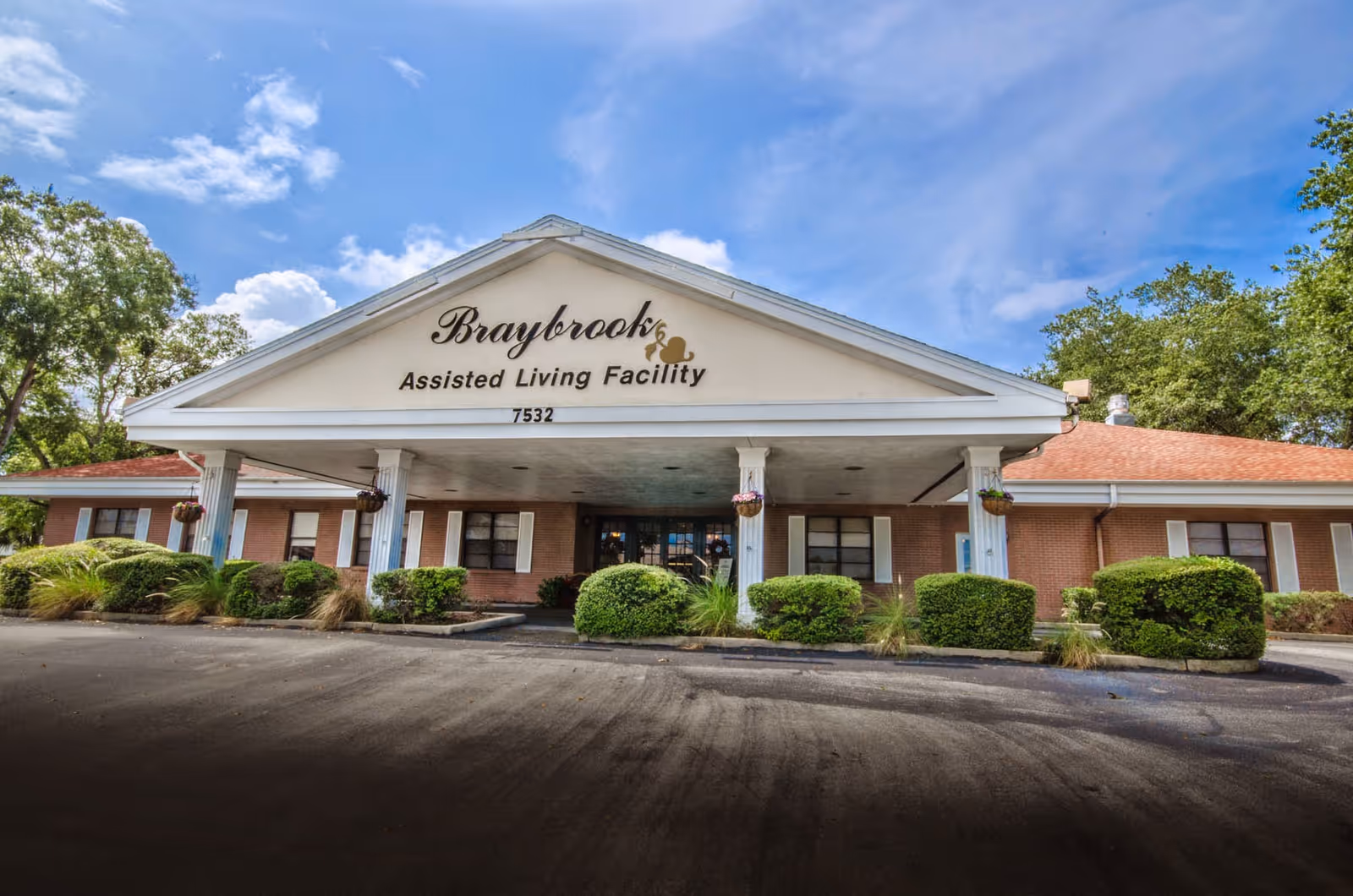 Front exterior view of Braybrook Assisted Living Facility building with a covered entrance supported by white columns, surrounded by bushes and trees under a partly cloudy blue sky.