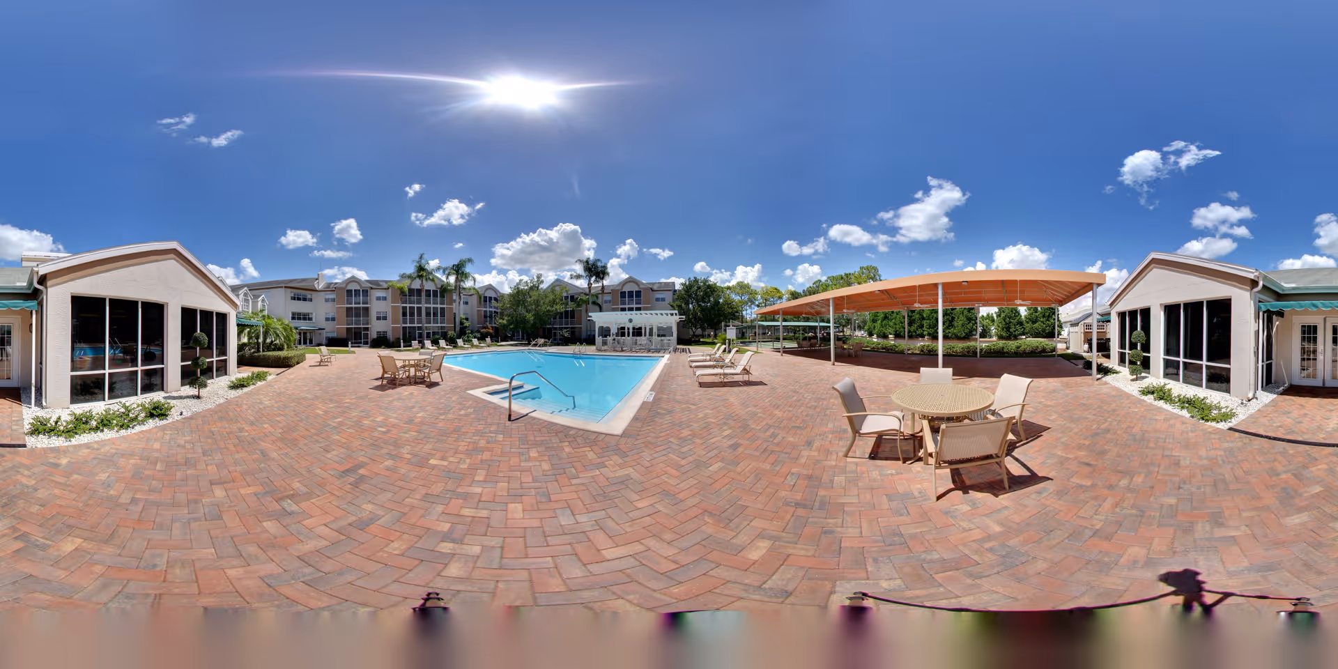 Outdoor patio area at Coral Oaks featuring a swimming pool surrounded by brick pavers, several lounge chairs, round tables with chairs, two buildings with large windows, and a covered seating area under a canopy. The sky is clear with a few clouds and the sun shining brightly.