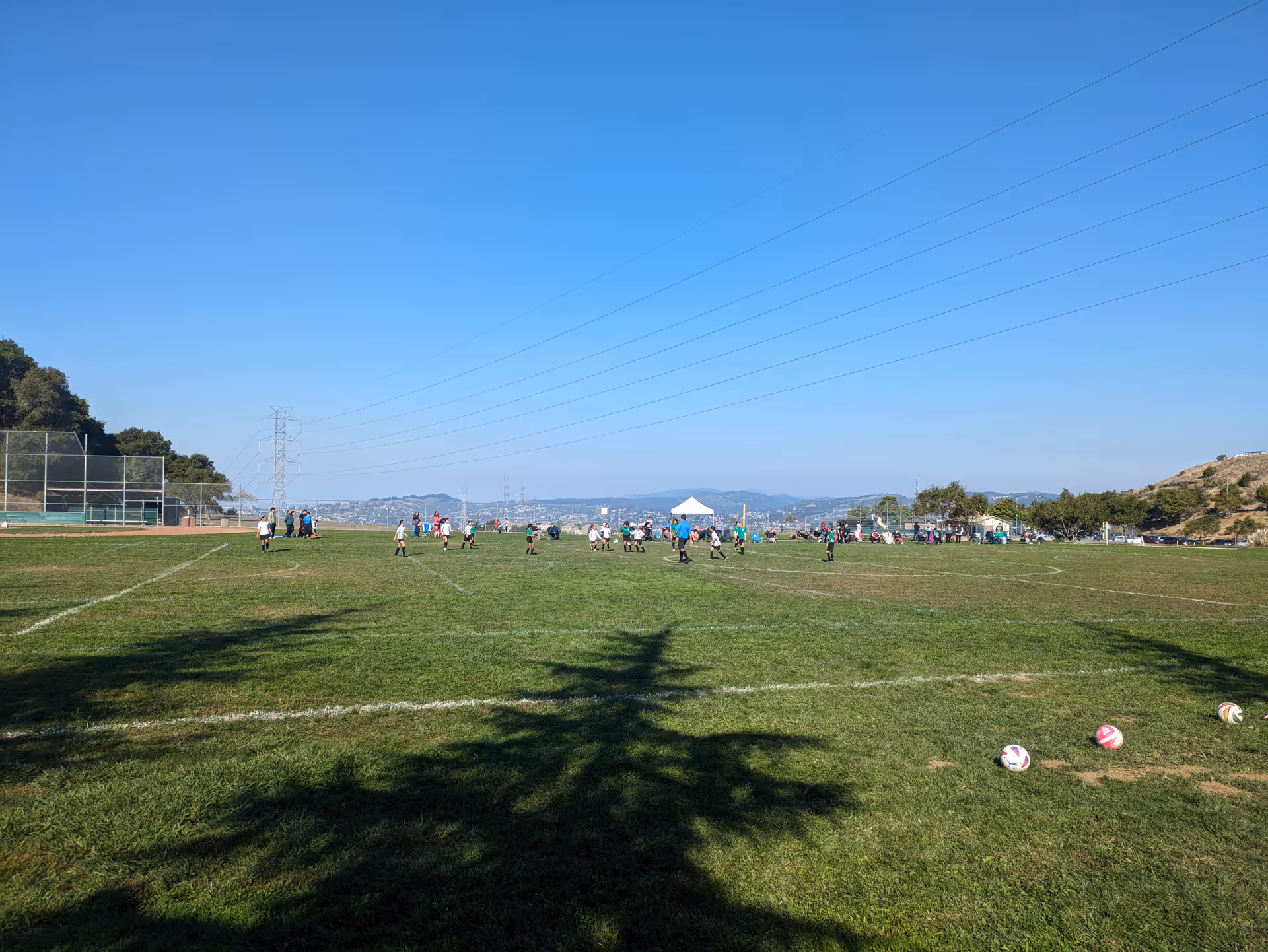 People playing soccer on a large sunny outdoor field with balls in foreground.