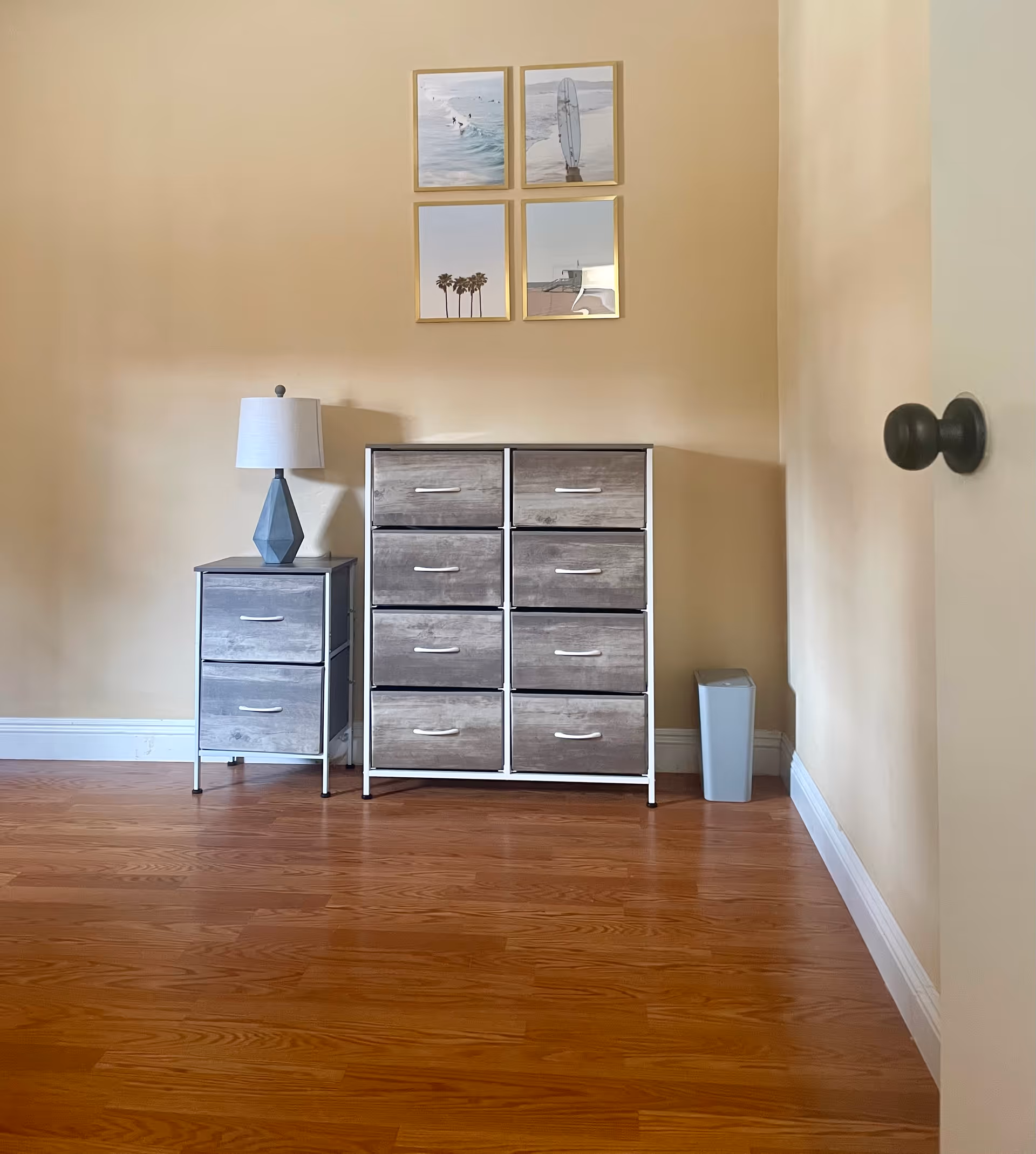 Corner of a beige bedroom with a lamp, a small two-drawer nightstand, and a larger dresser.