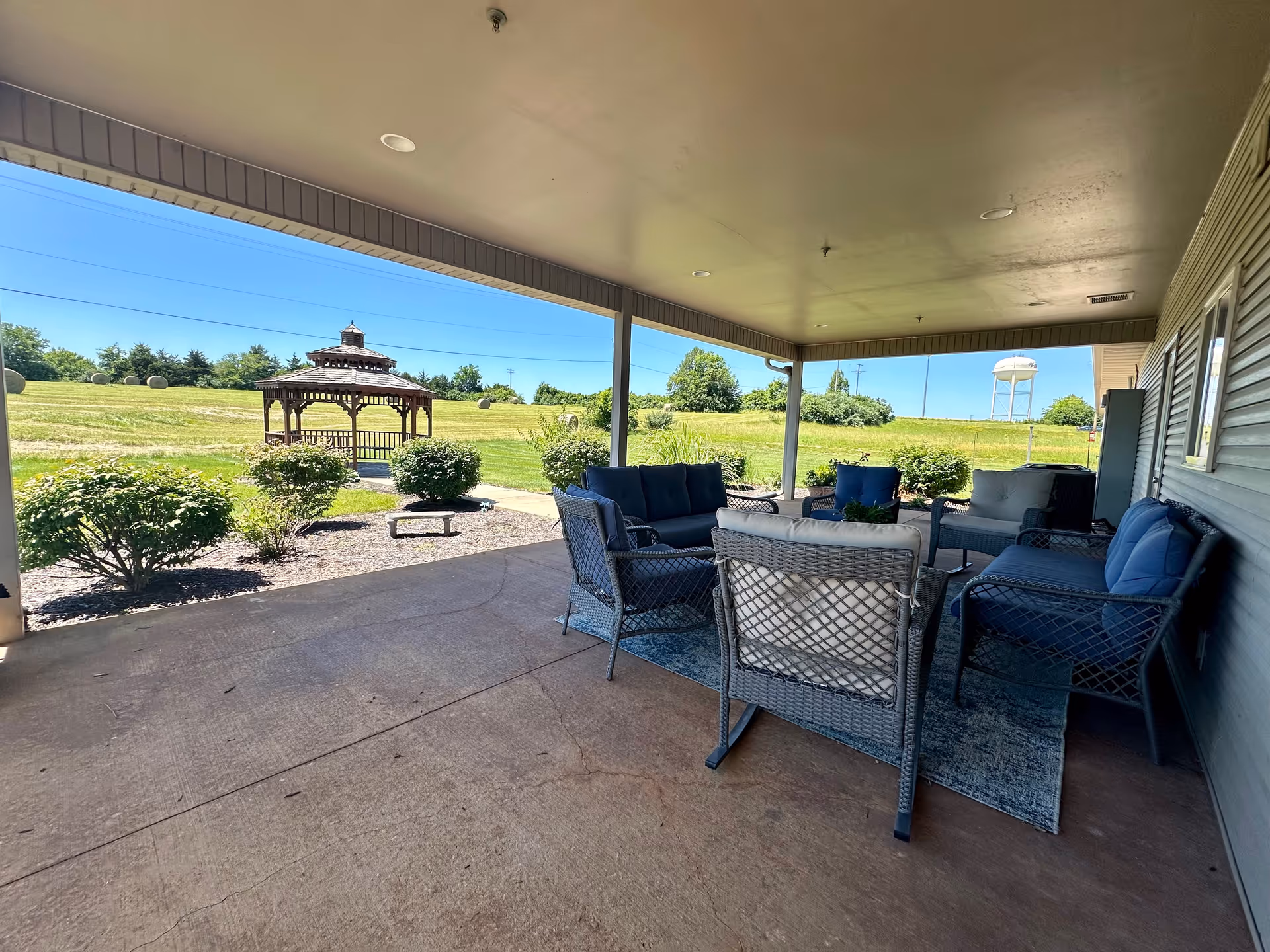 Covered outdoor patio area with cushioned wicker chairs and sofas arranged around a small table on a rug. Beyond the patio, there is a grassy area with bushes and a wooden gazebo. A water tower and clear blue sky are visible in the background.