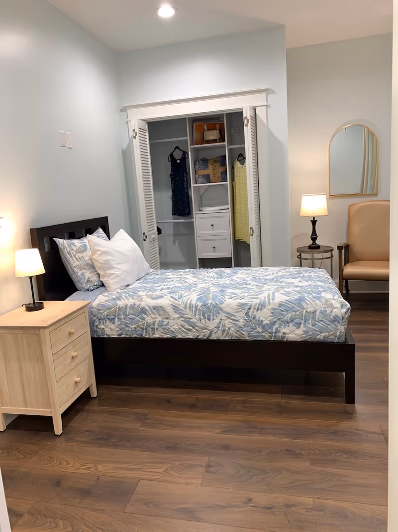 A tidy bedroom with a bed, nightstand, open closet, and seating in a sunlit pastel-hued room.