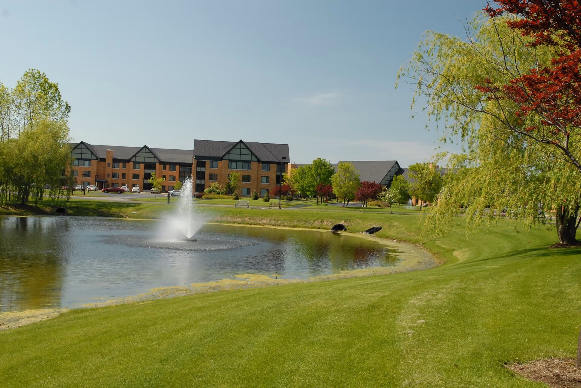 View of a large pond with a water fountain in the center, surrounded by green grass and trees with a multi-story brick building in the background under a clear blue sky.