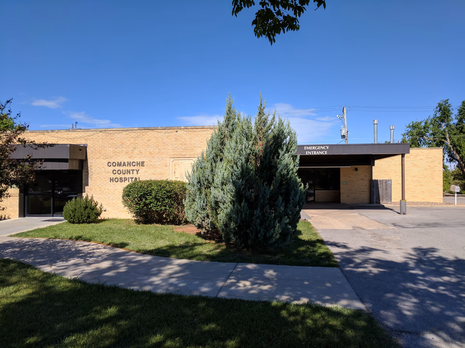 Brick hospital building with emergency entrance and surrounding trees.