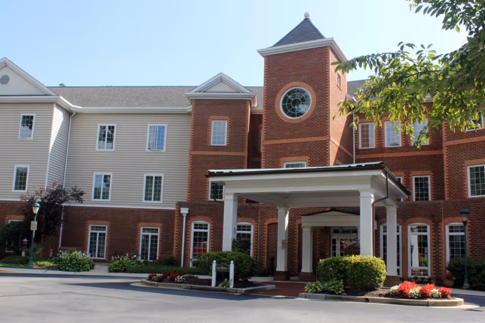 Front entrance of a brick and siding senior living building with a covered porte-cochère, tower with a round window, and landscaped driveway.