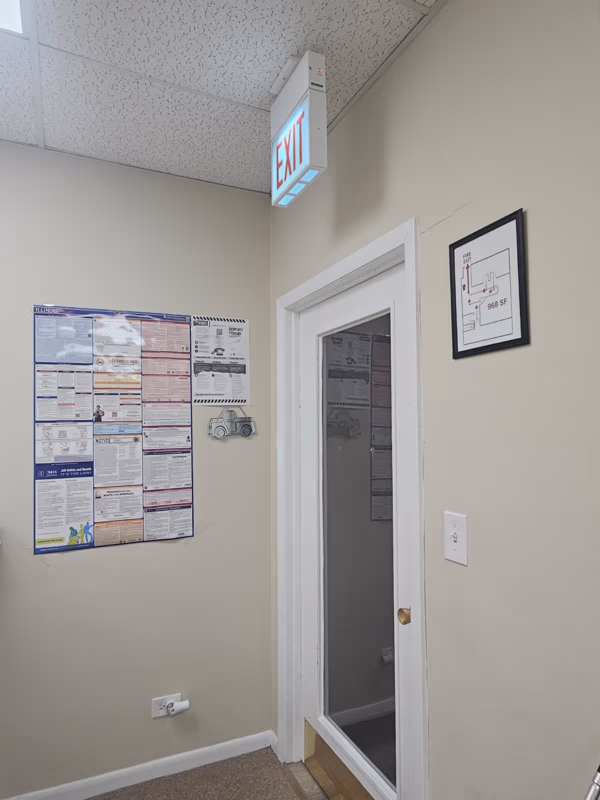 Interior hallway with an illuminated EXIT sign, posters on the wall, and a glass-paneled door.