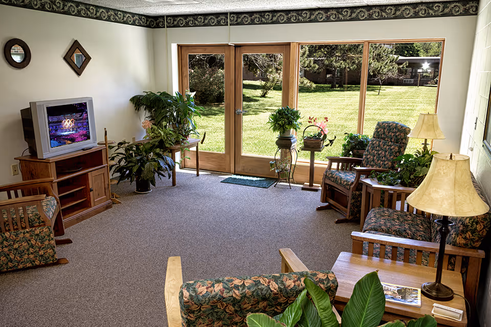 Sunlit common living room with floral-upholstered wooden chairs, a TV, potted plants, lamps, and large glass doors opening to a grassy courtyard.