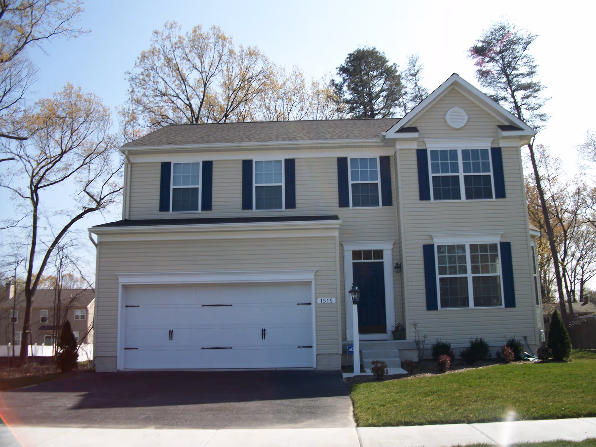 Two-story beige house with dark blue shutters and a two-car garage, surrounded by trees and a green lawn under a clear sky.