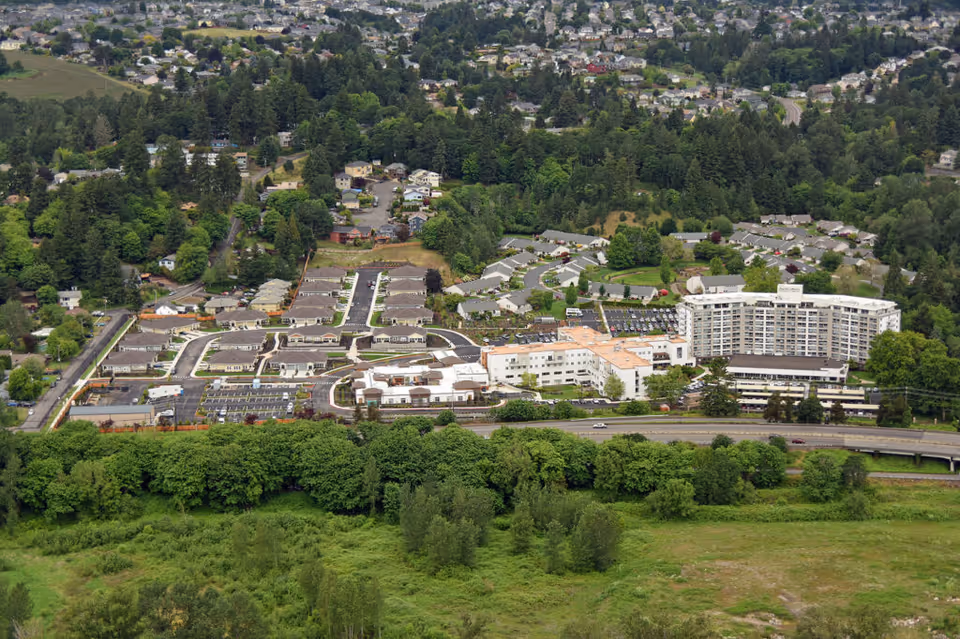 Aerial view of Capital Manor Retirement Community showing multiple buildings, parking lots, roads, and surrounding greenery with a residential neighborhood in the background.