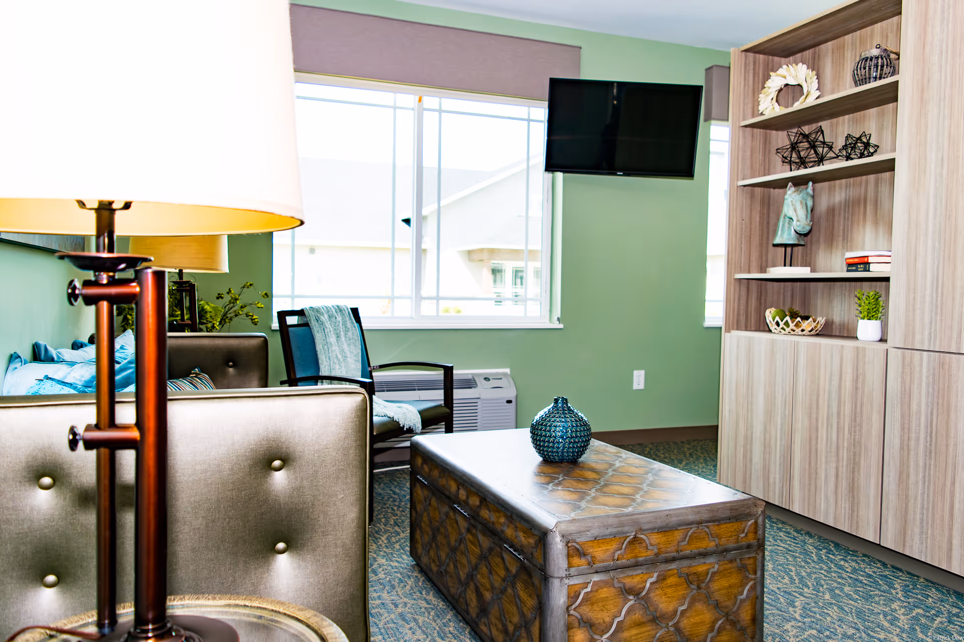A cozy living room area with a green wall, a window with a view of neighboring buildings, a mounted flat-screen TV, a wooden chest used as a coffee table with a decorative blue vase on top, a cushioned chair with a throw blanket, a floor lamp with a white shade, and a wooden shelving unit with various decorative items including a horse head sculpture, books, and plants.