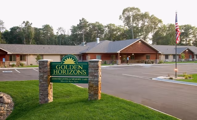 Exterior view of Golden Horizons assisted living and memory care facility with a large green sign in front, a paved parking lot, and a building surrounded by trees under a clear sky.