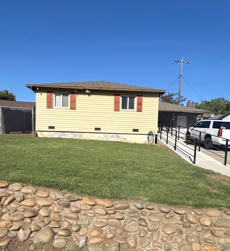 A single-story building exterior with beige siding, red shutters, a ramp, and a parked white SUV.