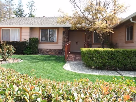 Single-story brown senior living home with central entrance, trimmed hedges, and a green lawn.