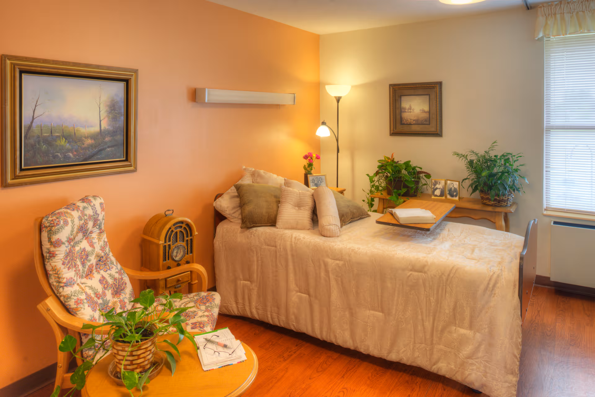 A cozy bedroom in a senior living facility with a bed, plants, and seating.