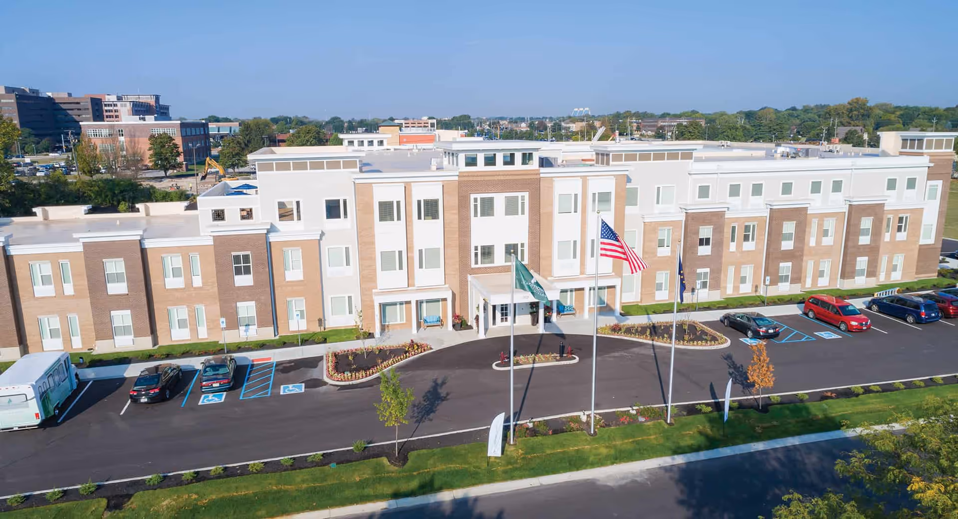 Aerial view of the front of a three-story brick-and-white senior living building with flagpoles, a landscaped entrance, and parked cars.