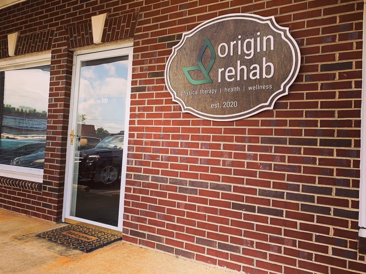 Brick exterior of Origin Rehab facility with glass door and large oval sign.
