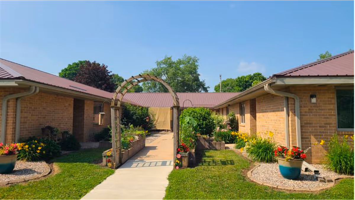 Outdoor garden area at Meadow View Assisted Living & Memory Care with a concrete walkway leading through a wooden archway surrounded by flower beds and potted plants, flanked by single-story brick buildings with red metal roofs under a clear blue sky.