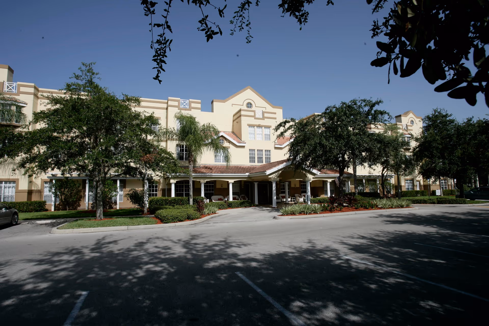 Exterior view of a multi-story senior living facility with beige walls and a tiled roof, surrounded by trees and landscaping, with a driveway and parking area in front.