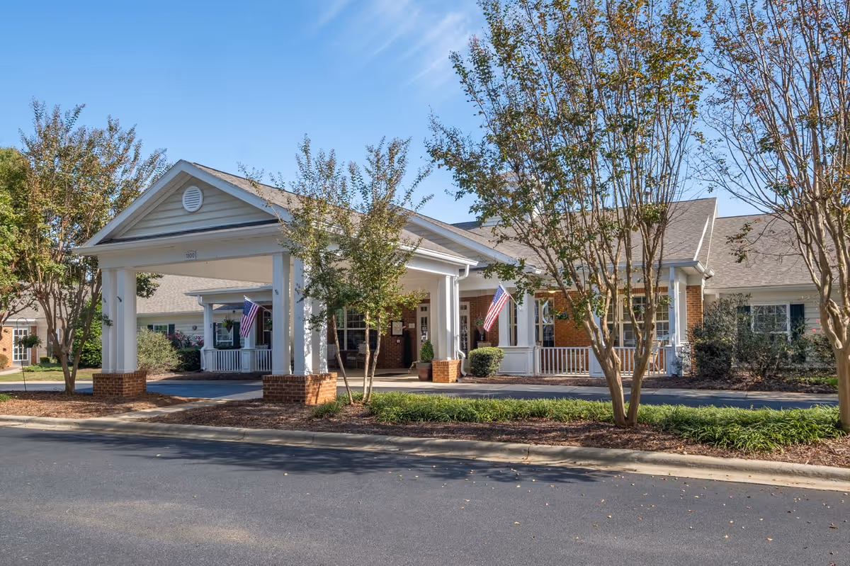Front exterior view of Chandler Place Assisted Living and Memory Care building with a covered entrance supported by white columns and brick bases, American flags displayed, surrounded by trees and landscaping under a clear blue sky.