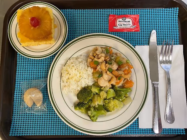 A meal tray with a plate containing white rice, cooked broccoli, and a stir-fry dish with cashews and vegetables. There is a small plate with a pineapple upside-down cake topped with a cherry, a wrapped fortune cookie, a packet of Kikkoman soy sauce, and a knife and fork placed on a white napkin. The tray has a blue placemat underneath the dishes.