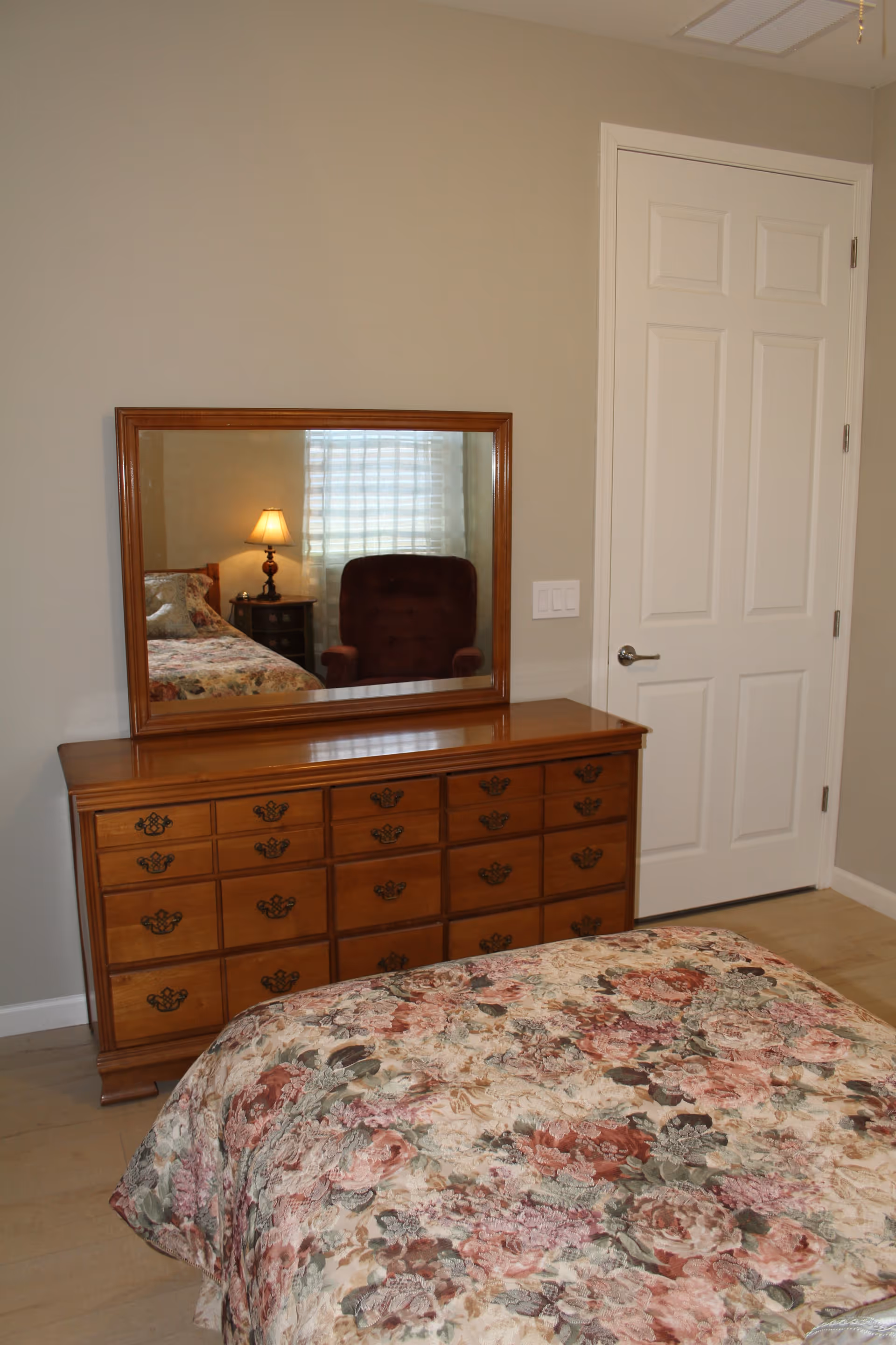 A bedroom with a wooden dresser and mirror reflecting a lamp and chair, and a floral bedspread in the foreground.