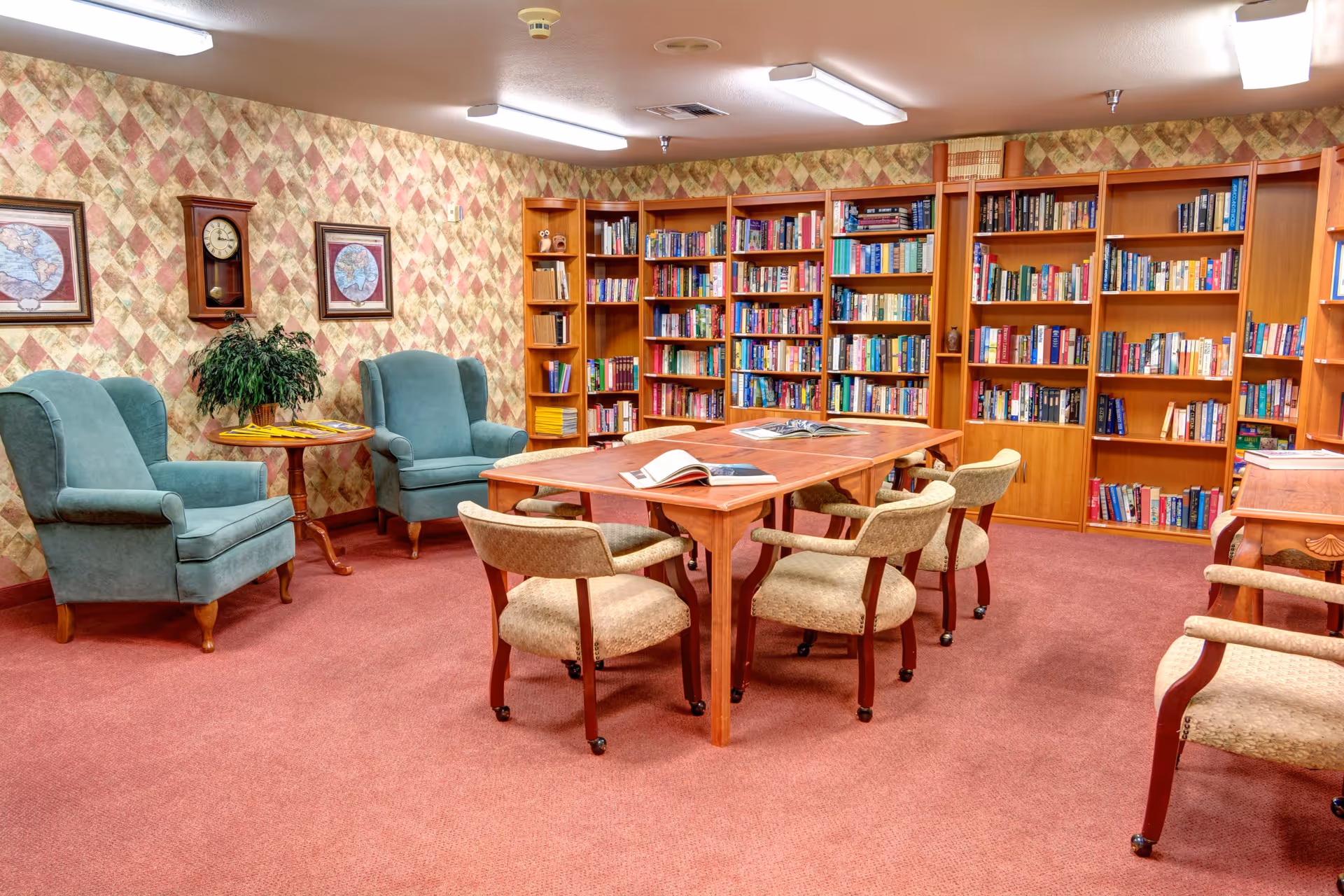 A cozy library room with wooden bookshelves filled with books along the walls, a wooden table in the center with open books on it, surrounded by cushioned chairs with wooden arms. Two teal armchairs and a small round table with a plant are placed against a patterned wallpapered wall with framed maps and a wall clock.
