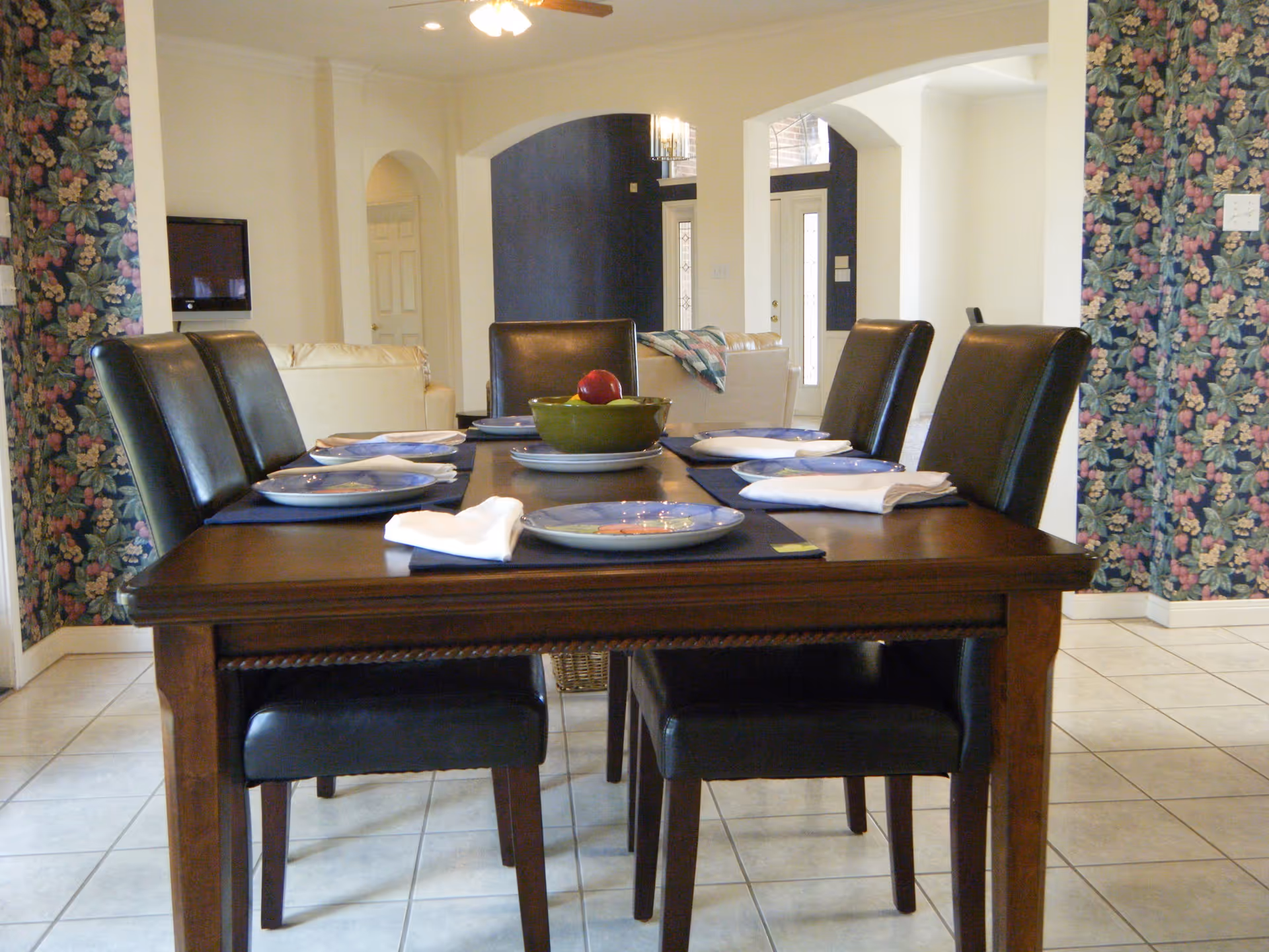 A wooden dining table set for six with plates in a floral-walled dining room.